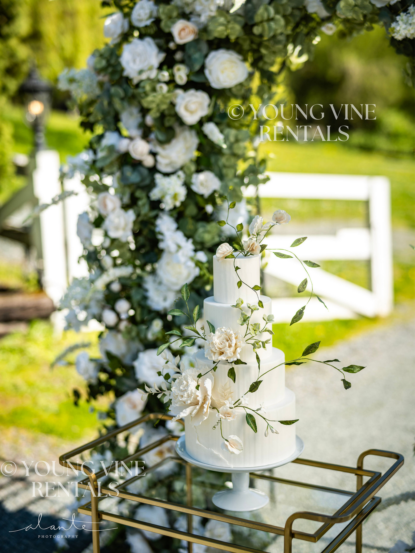 Wedding cake with ivory and green sugar flowers displayed on a gold bar cart, in front of a lush floral arbor at an outdoor winery wedding. The wedding arch in the background is studded with an abundance of ivory and white flowers and various types of eucalyptus foliage.