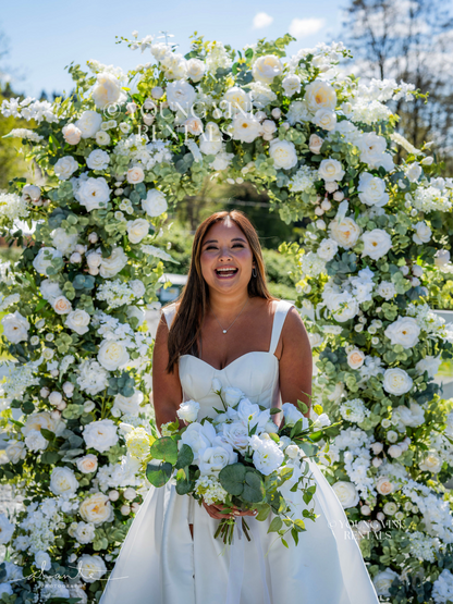 Outdoor winery wedding ceremony with a tan brunette bride standing in front of a lush floral arbor, wearing a modern white satin ballgown. She is holding a white and green floral bouquet with a loose organic shape. The arbor behind her is lush and romantic, and is studded with an abundance of ivory and white flowers and various types of eucalyptus foliage.