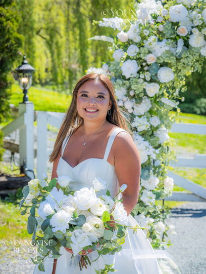 Outdoor winery wedding ceremony with a smiling, tan, brunette bride standing in front of a lush floral arbor, wearing a modern white satin ballgown. She is holding a white and green floral bouquet with a loose organic shape. The arbor behind her is lush and romantic, and is studded with an abundance of ivory and white flowers and various types of eucalyptus foliage.