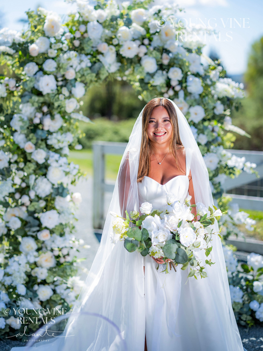 Outdoor winery wedding ceremony with a smiling, tan, brunette bride standing in front of a lush floral arbor, wearing a modern white satin ballgown and a cathedral veil. She is holding a white and green floral bouquet with a loose organic shape. The arbor behind her is lush and romantic, and is studded with an abundance of ivory and white flowers and various types of eucalyptus foliage.