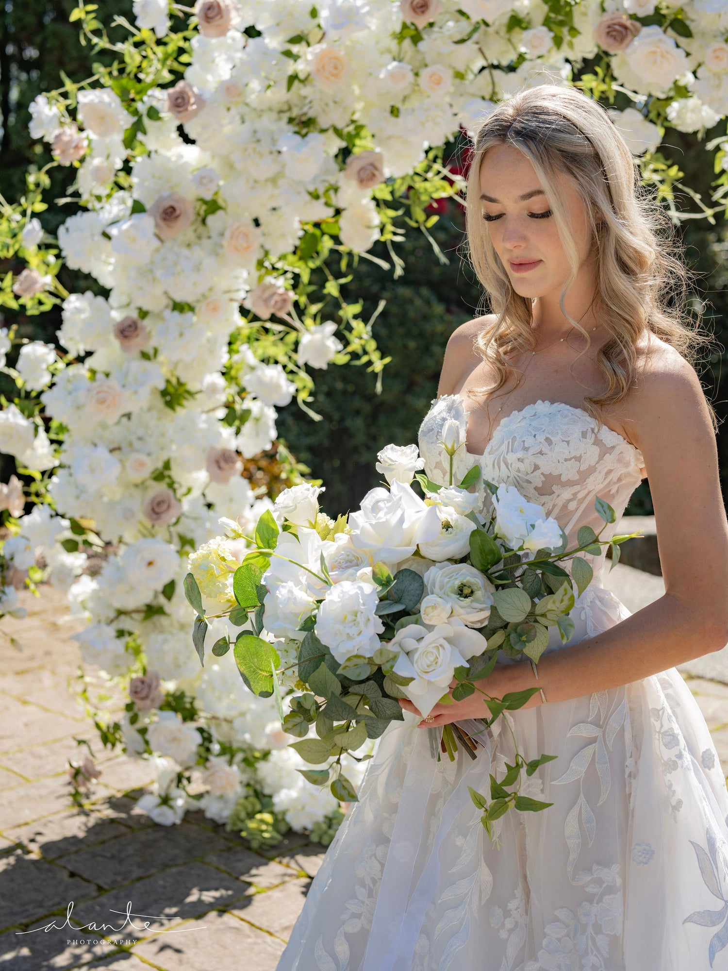 Bride standing in front of wedding ceremony arch packed with flowers in white, ivory, taupe, and pale blush, accented by delicate vines.